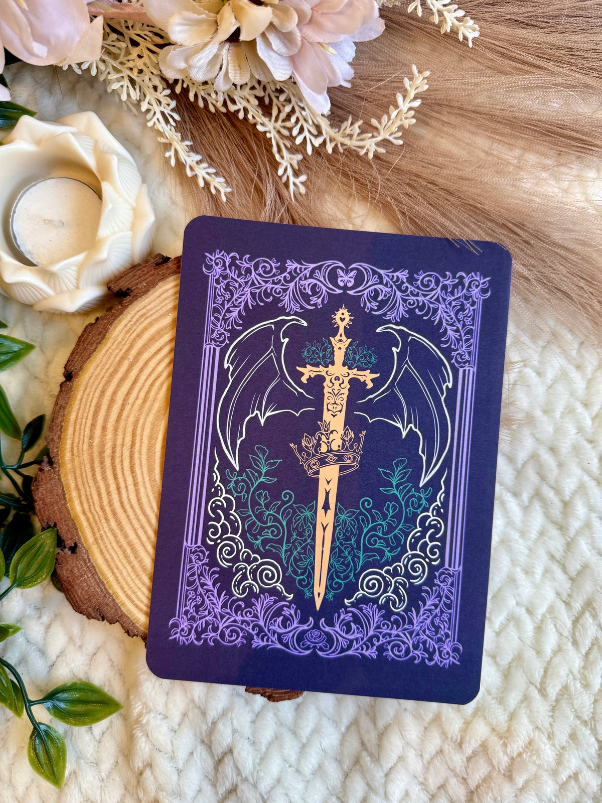 A kindle insert with a dark purple cover featuring a gold winged sword and silver filigree, styled on a cream knit blanket with a wood slice, dried flowers, and a candle.