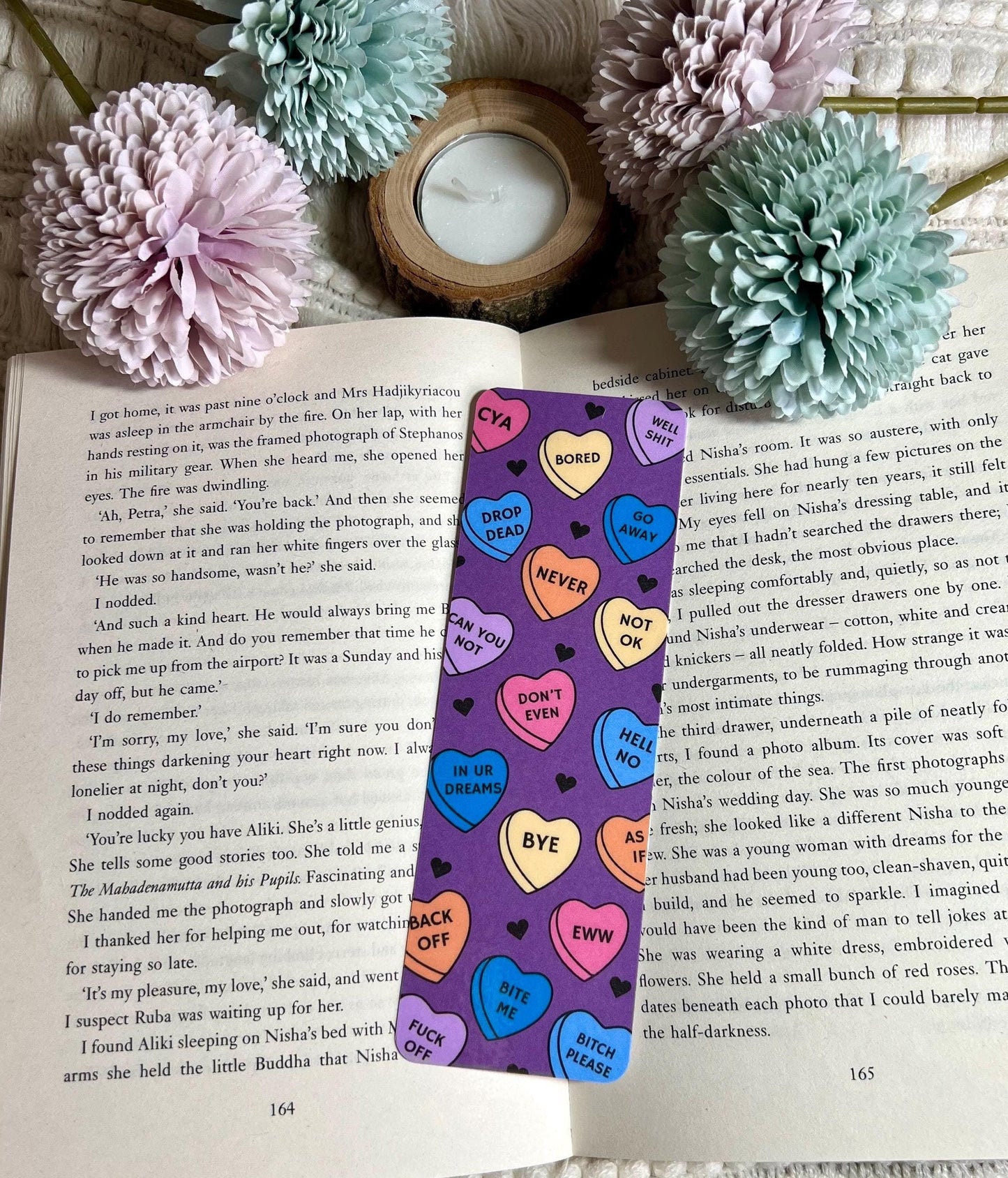 A bookmark with a sarcastic heart design, placed on an open book, with decorative flowers and a candle in the background.
