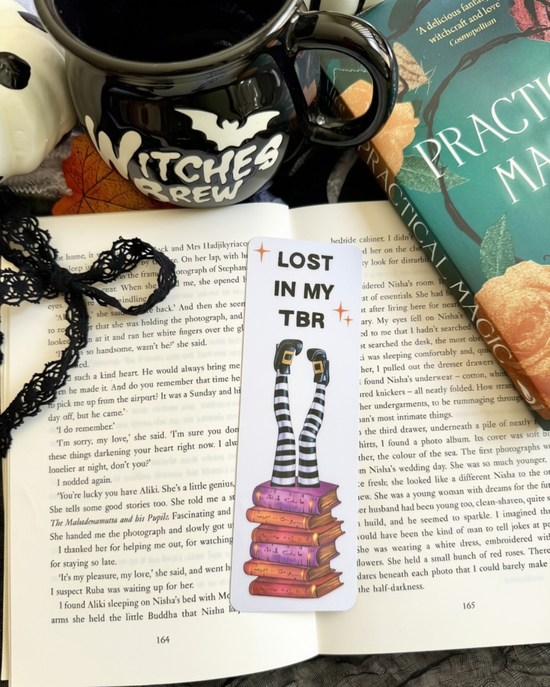 A bookmark of a witches legs stuck in a pile of books with the text, lost in my tbr. The bookmark is placed on an open book surrounded with Halloween decorations. 