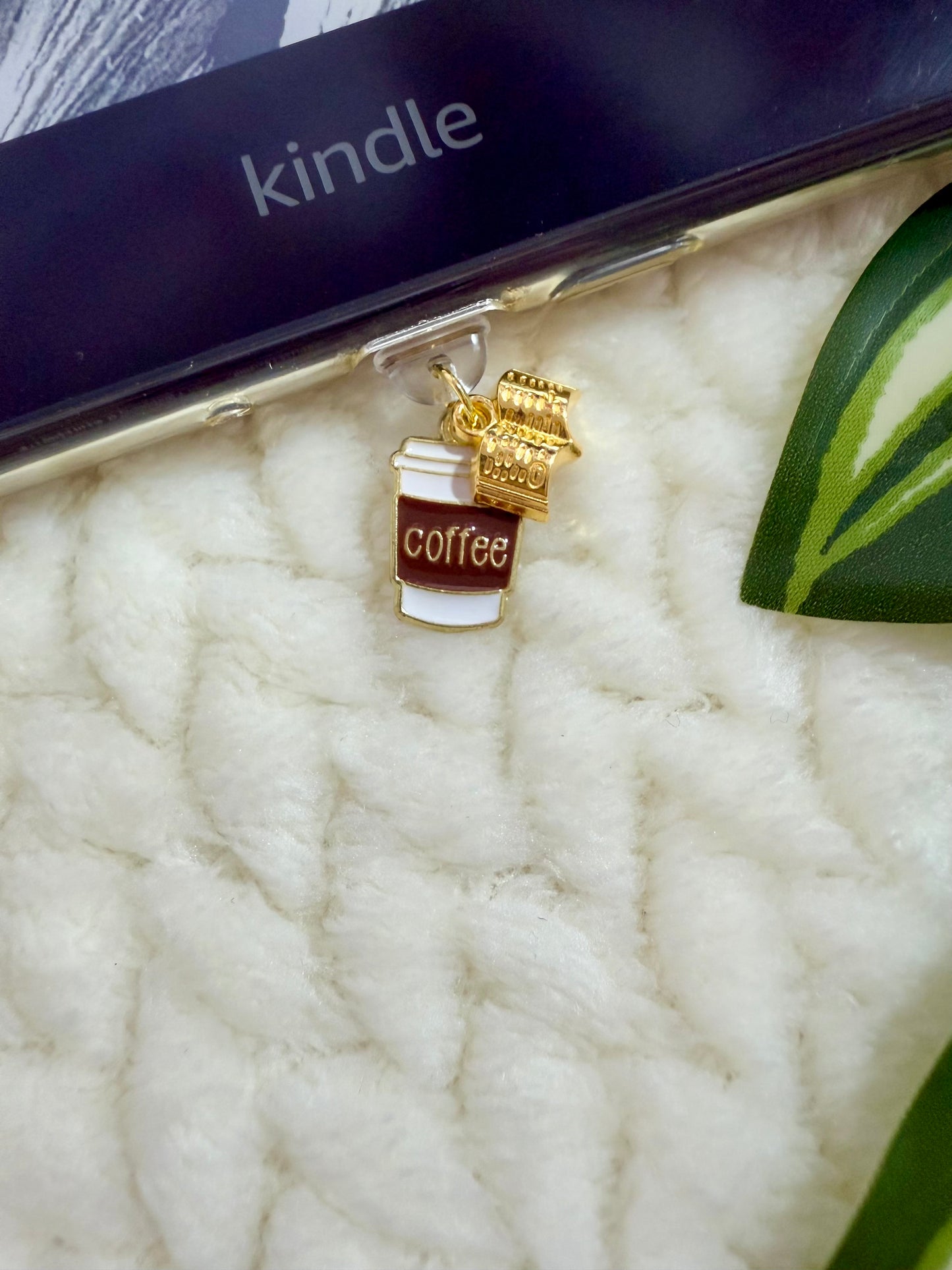 Gold coffee cup and book charm on a white textured surface with a Kindle device in the background.