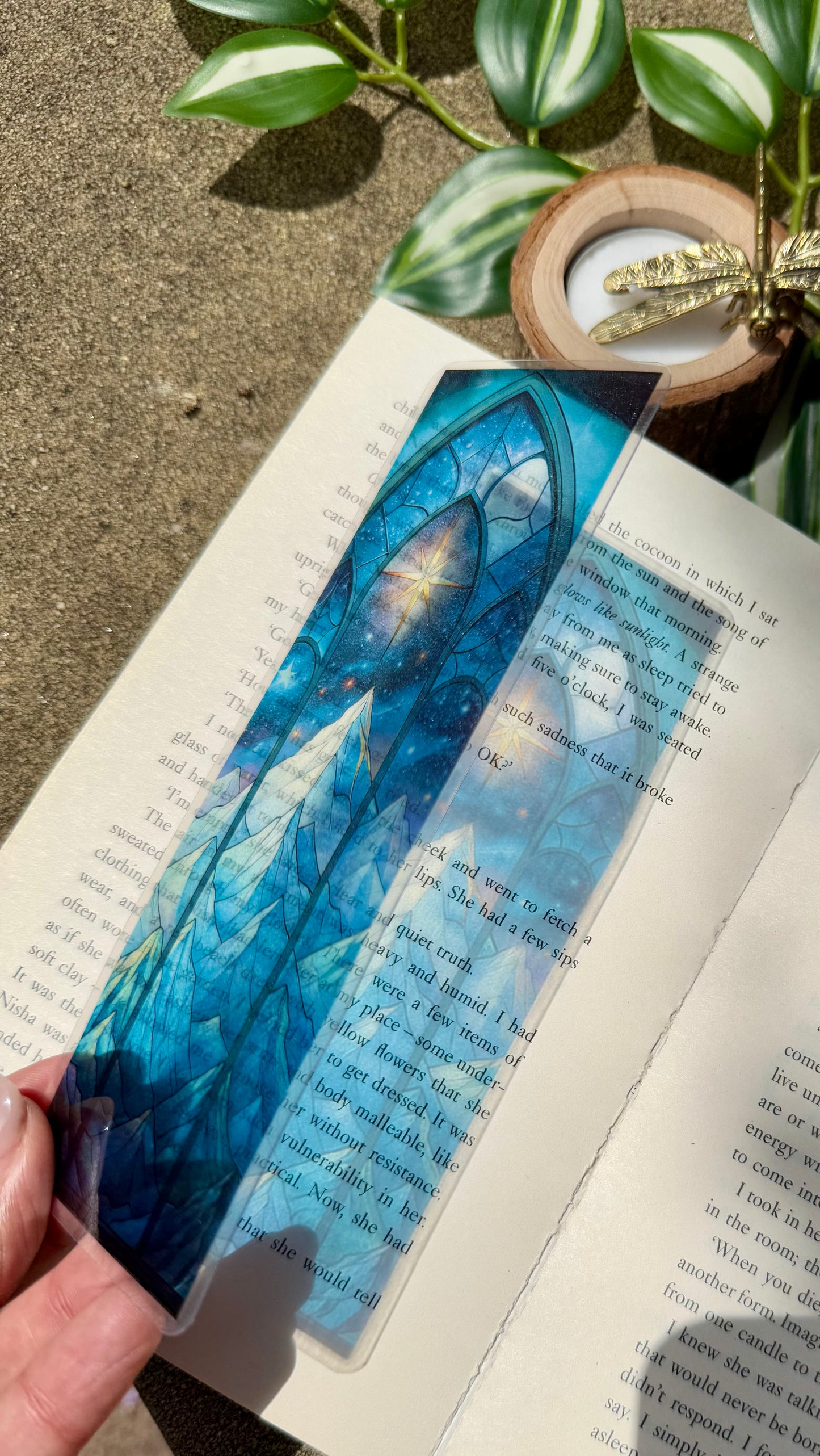 A stained glass bookmark of a mountain and night stars. The bookmark is placed over an open book with the reflection of the bookmark showing on the open book.