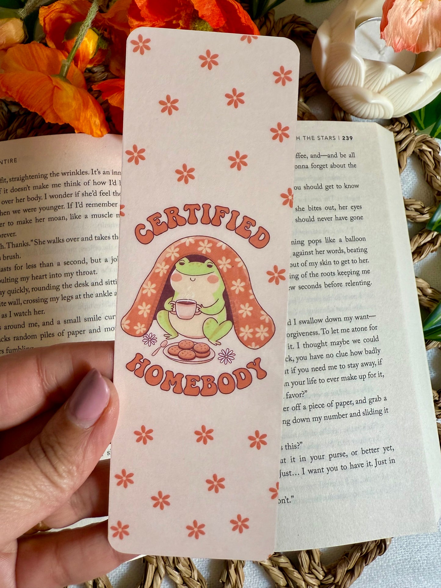 Hand holding a bookmark of a frog illustration and text saying 'Certified Homebody' on a book page.