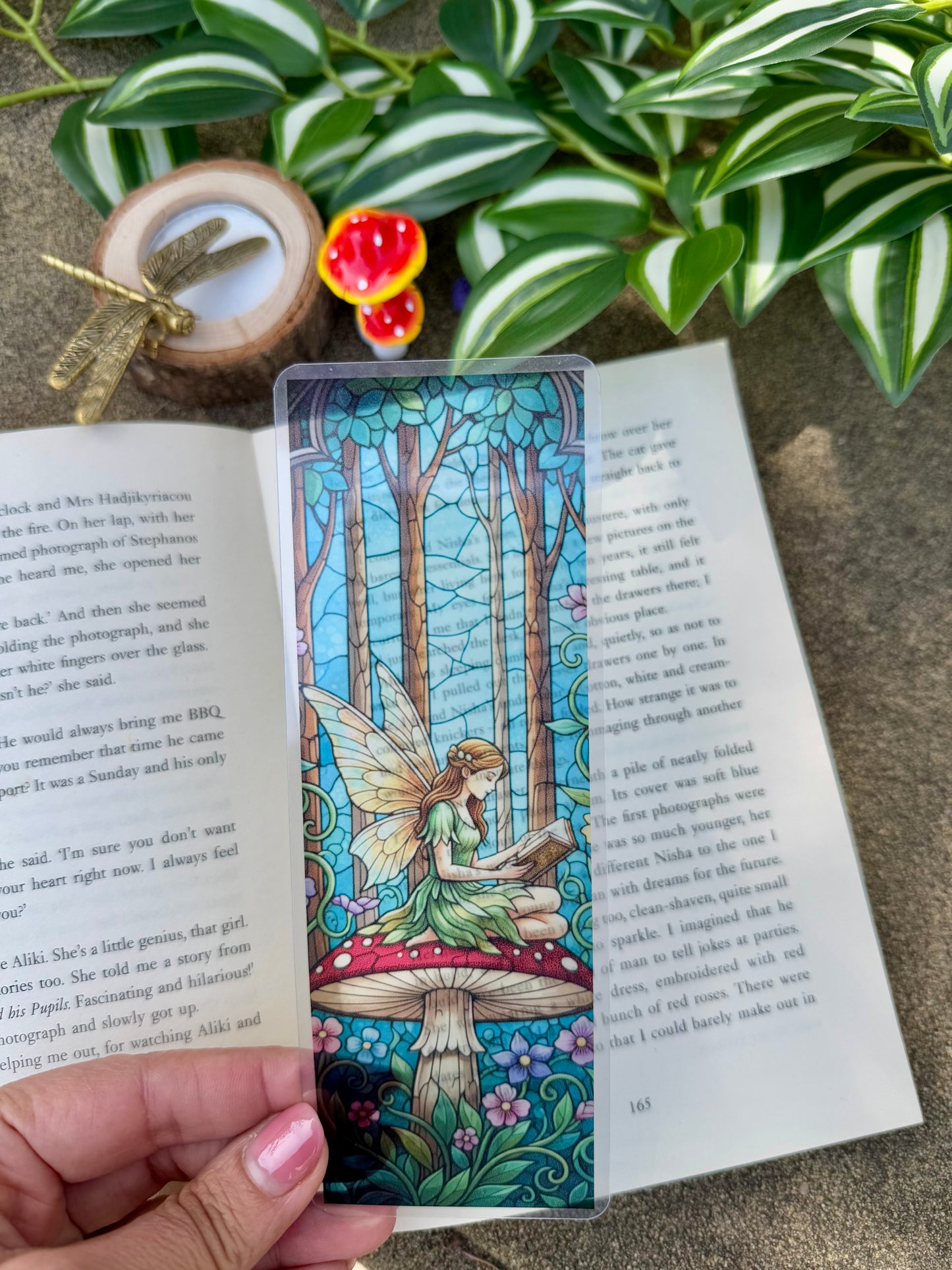 A transparent stained glass bookmark of a fairy sitting on a mushroom in a forest reading a book.