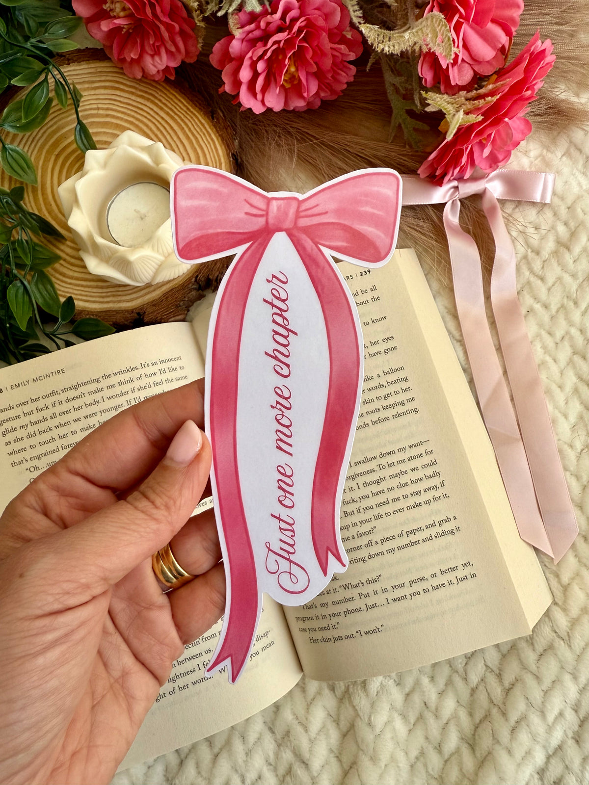 A hand holds a pink paper bookmark shaped like a bow with the text 'Just one more chapter' printed on it, positioned over an open book. The scene is styled with pink flowers, a woven tray, and a pink ribbon on a cream knit blanket.