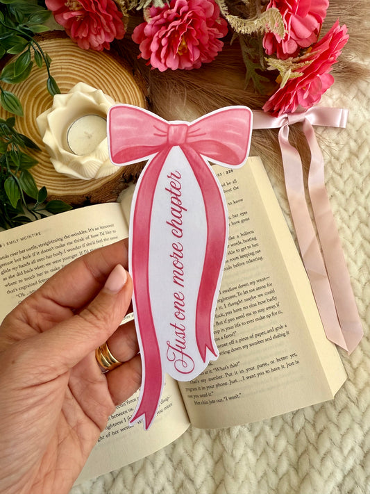 A hand holds a pink paper bookmark shaped like a bow with the text 'Just one more chapter' printed on it, positioned over an open book. The scene is styled with pink flowers, a woven tray, and a pink ribbon on a cream knit blanket.