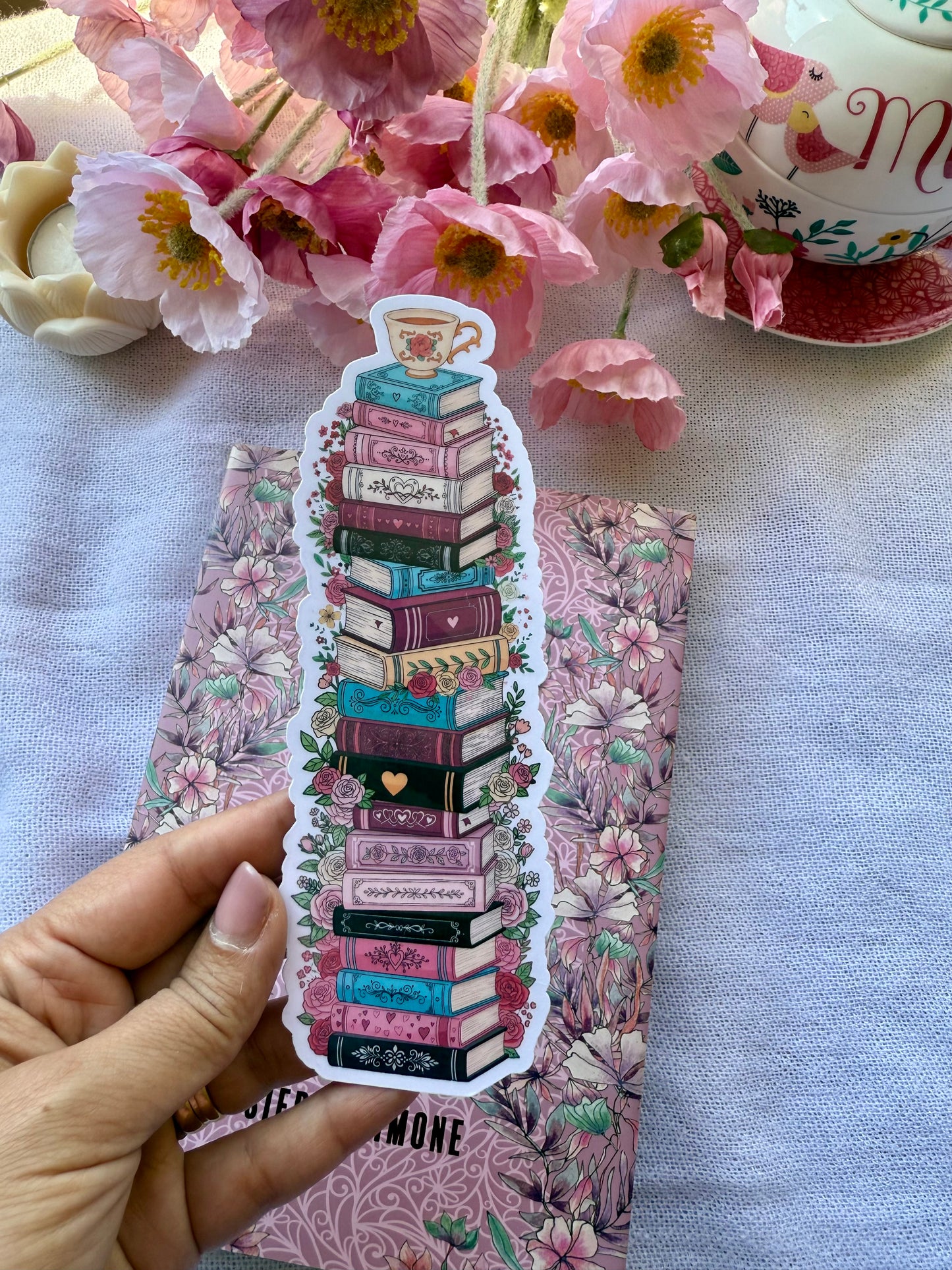 A die cut bookmark of a floral stack of books with a tea cup sitting on top of the books. The bookmark is held over a floral book with flowers and tea in the background.