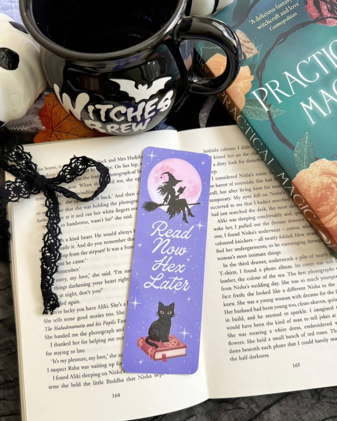 A purple witch-themed bookmark with a black cat silhouette and pink moon, reading 'Read Now Hex Later,' placed on open book pages surrounded by Halloween decor, a black witch hat, and a hand holding the page.