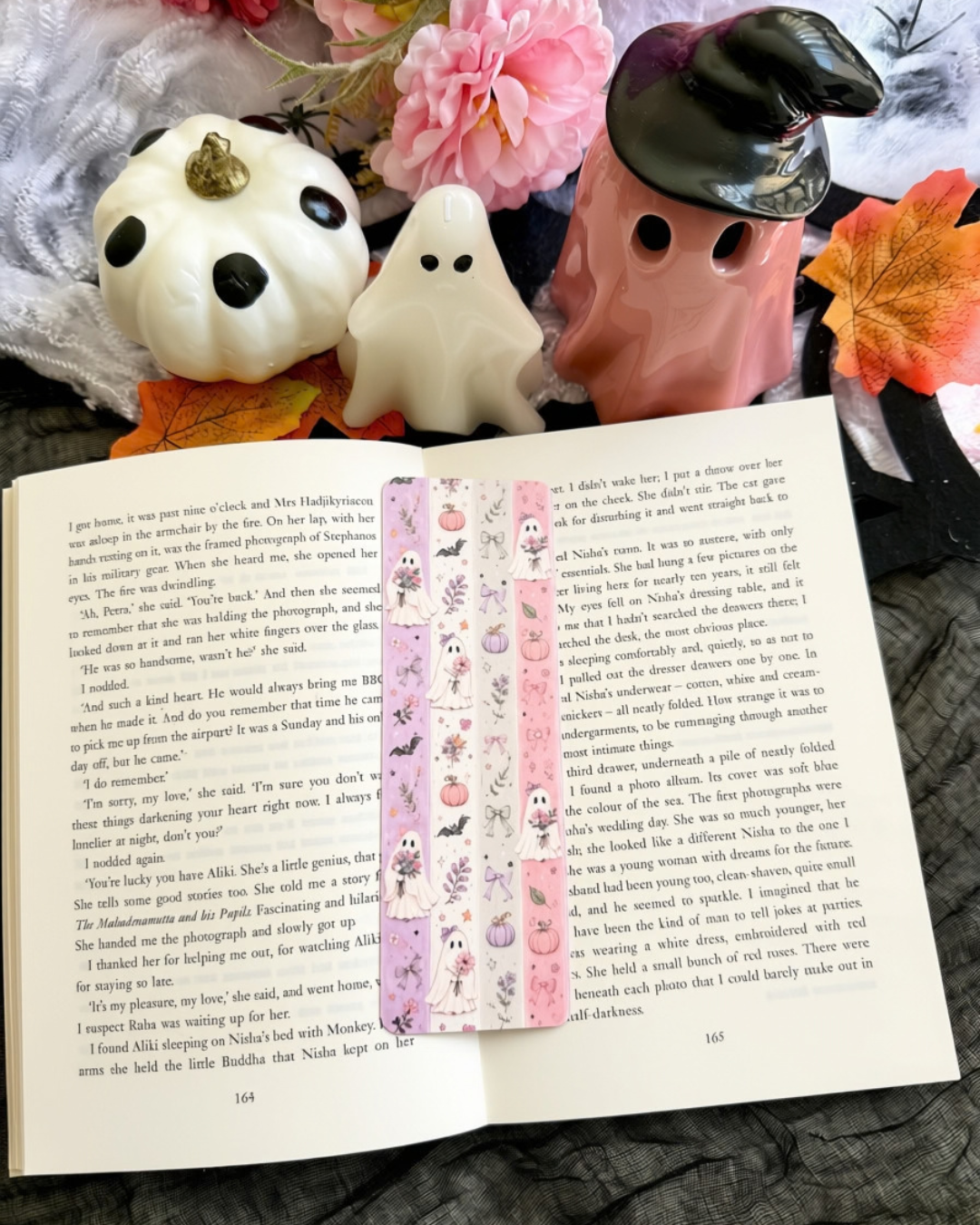 An open book spread with printed text and a vertical decorative bookmark featuring pink cartoon characters, styled on a bed with plush toys, flowers, and autumn leaves.