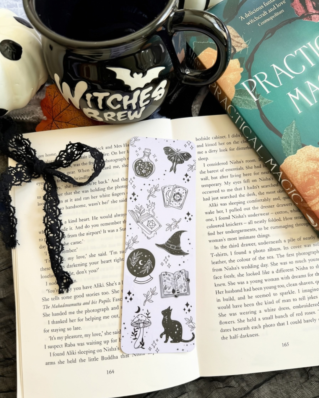 A hand holds a book titled 'The Practice of Witchcraft' while another book lies open underneath, decorated with black lace bookmarks featuring a witch illustration and a black cat design. The scene is styled with a black witch hat, a skull, and autumn leaves.