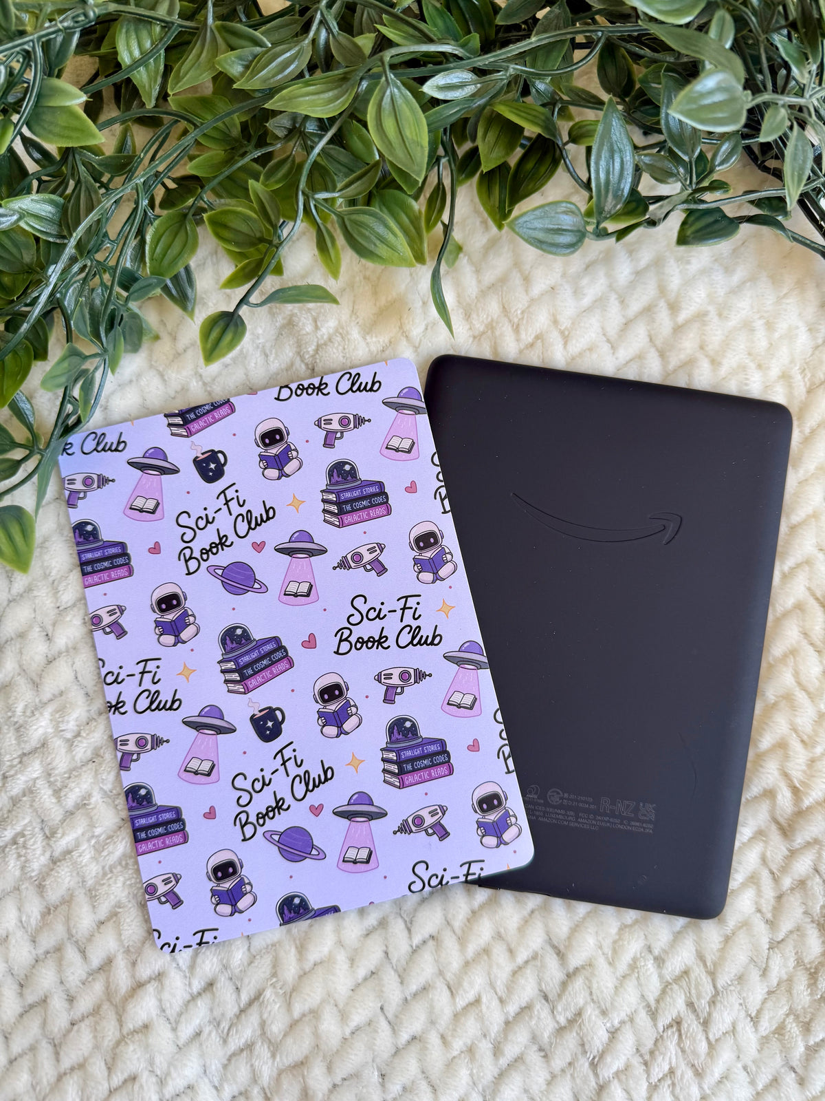 A purple kindle insert with a cartoon sci-fi astronaut pattern lies next to a black Kindle e-reader on a cream knit blanket with green leaves at the top.