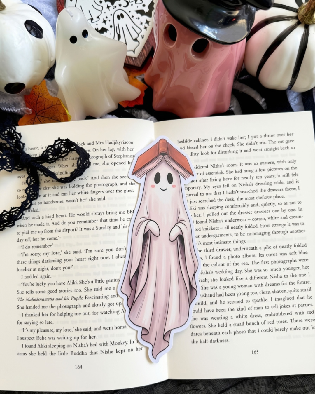 A die-cut bookmark of a ghost with an open book on its head. The bookmark is placed on an open book surrounded by Halloween decorations. 