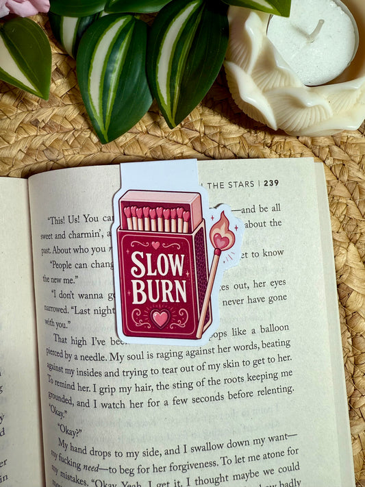 A rectangular magnetic bookmark shaped like a red matchbox with the words 'SLOW BURN' and a small flame icon, placed on an open book. The scene includes green leaves and a white candle in a woven mat background.