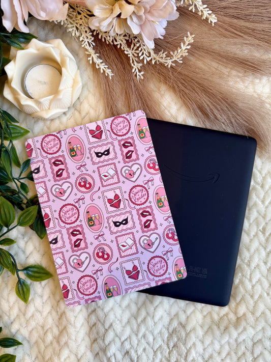 A pink notebook with a spicy themed pattern of hearts, lips, and masks, placed next to a black tablet on a cream knit blanket with decorative dried flowers and green leaves.