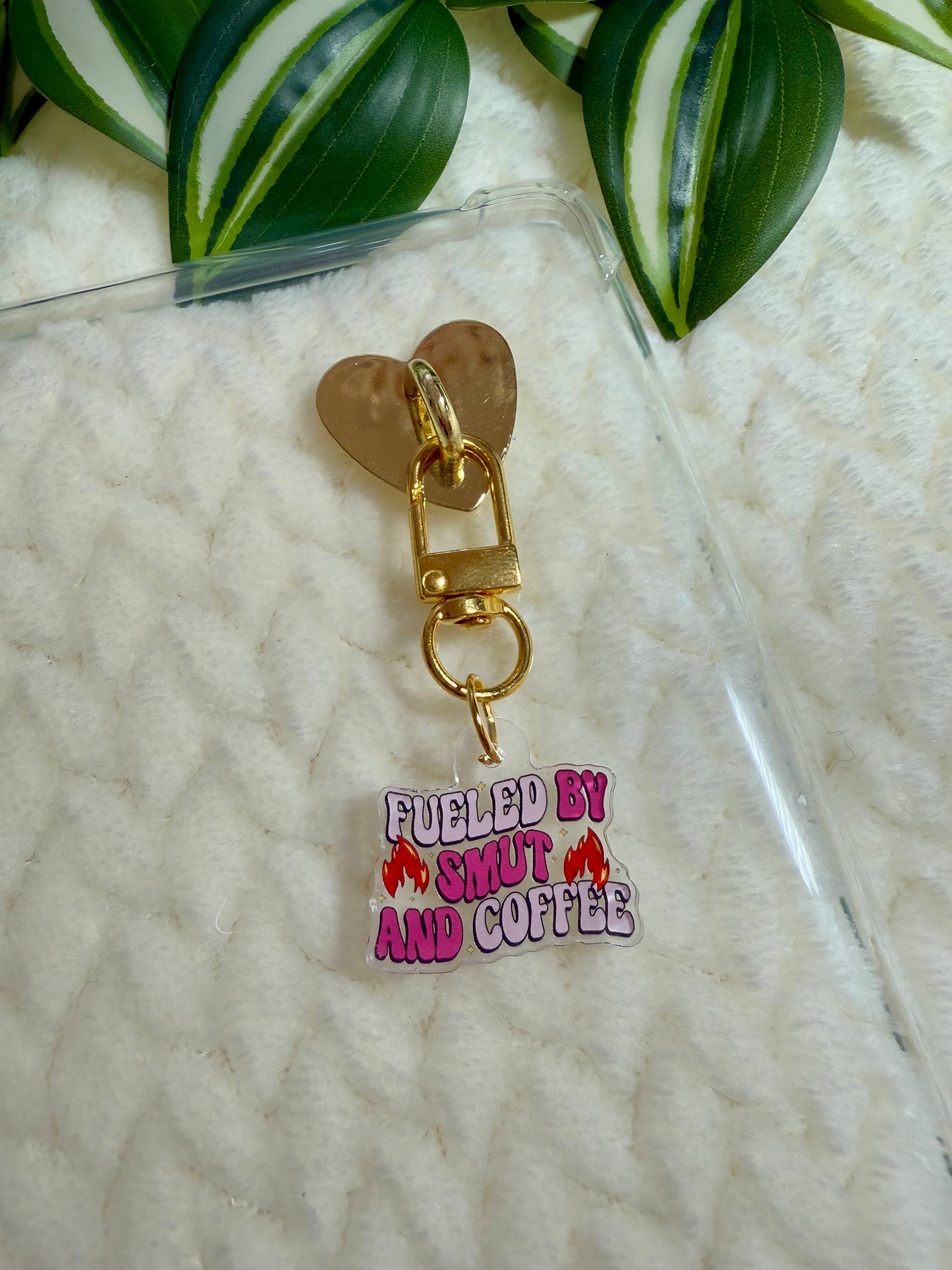 A charm with the text 'Fueled by Smut and Coffee' attached to a clear kindle case.