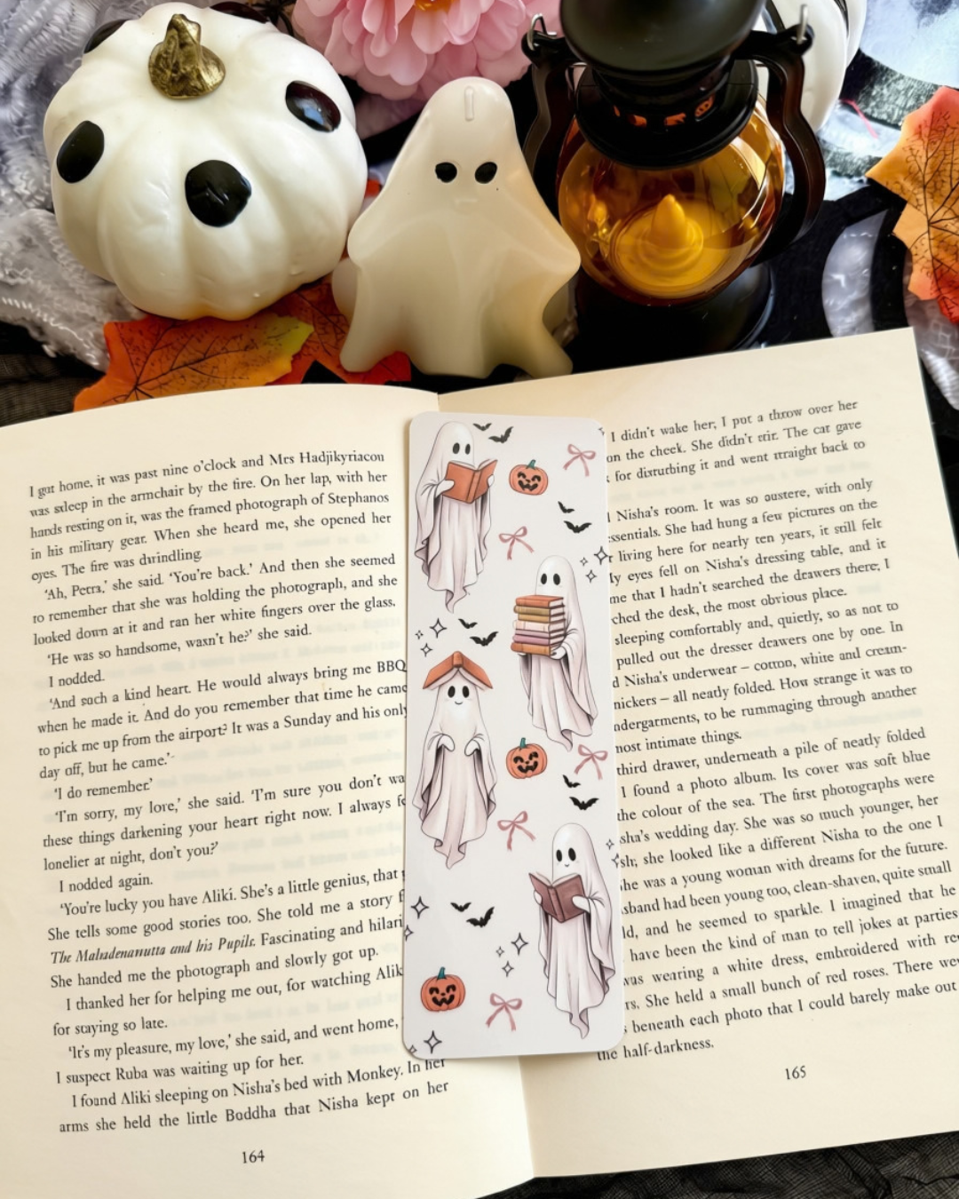 A bookmark of various ghosts reading books. The bookmark is place on an open book surrounded by Halloween decorations 
