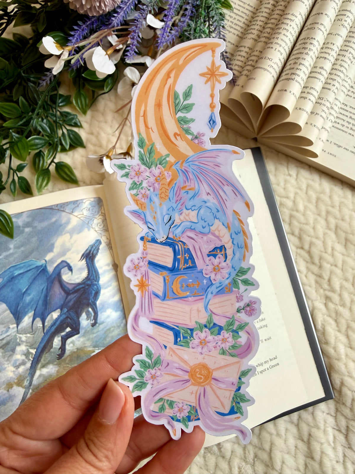 A die cut bookmark of a dreamy dragon on a set of books. the bookmark is placed over an open book depicting a page of a blue dragon
