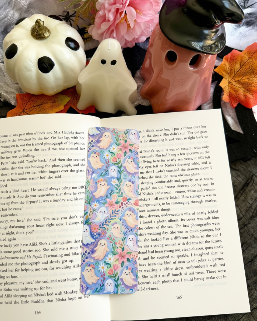 An open book spread with printed text and a colorful bookmark featuring a ghost and pastel flowers, styled on fabric with pink flowers and a small ghost figurine nearby.