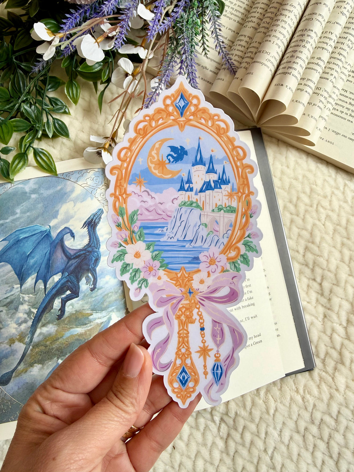 A die cut bookmark or a mirror with a fantasy scene of a castle and dragon. the bookmark is placed over a fantasy book depicting a blue dragon
