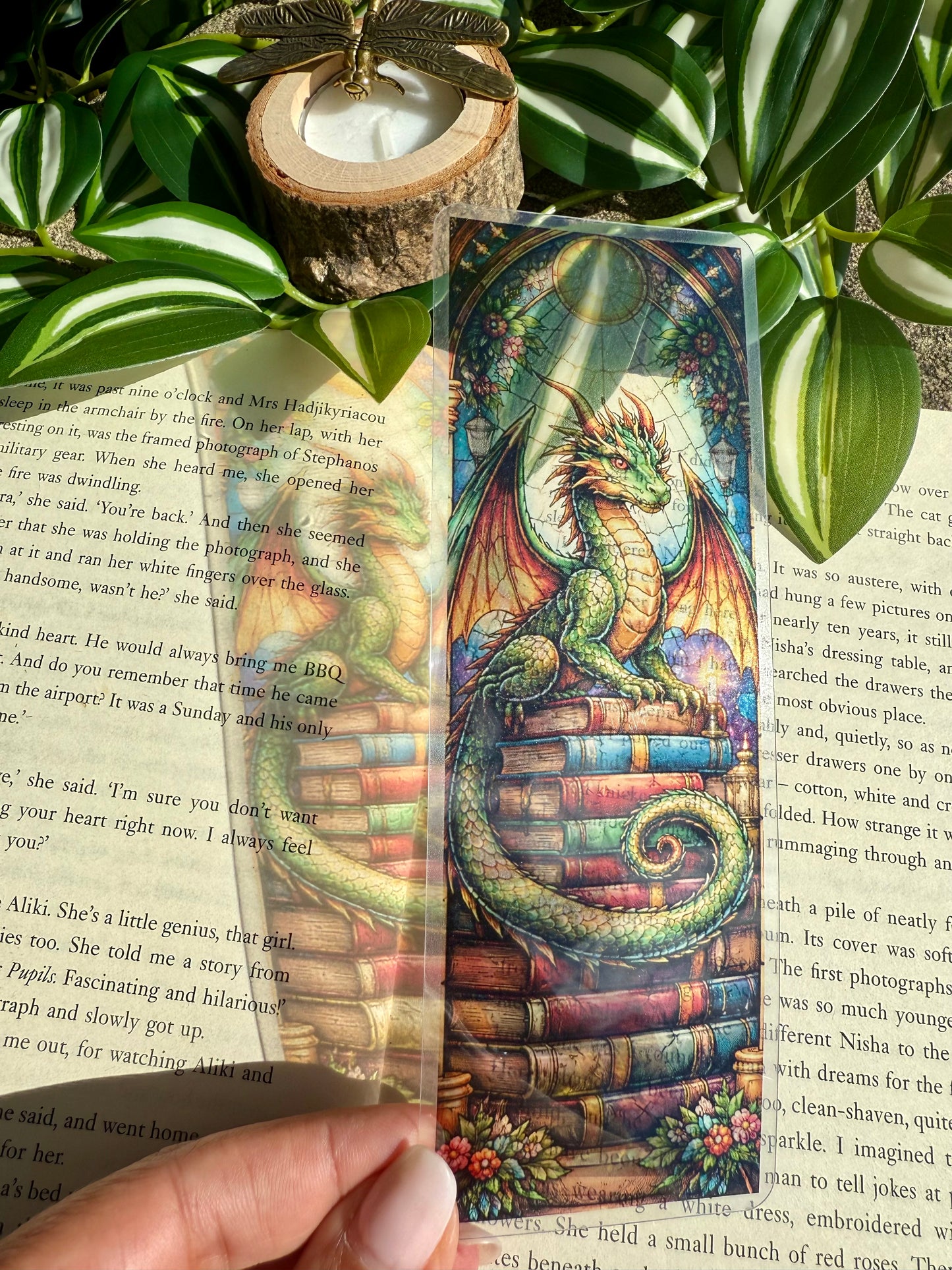 A colorful stained glass bookmark of a dragon sitting on a stack of books. The bookmark is held over a book with green leaves in the background