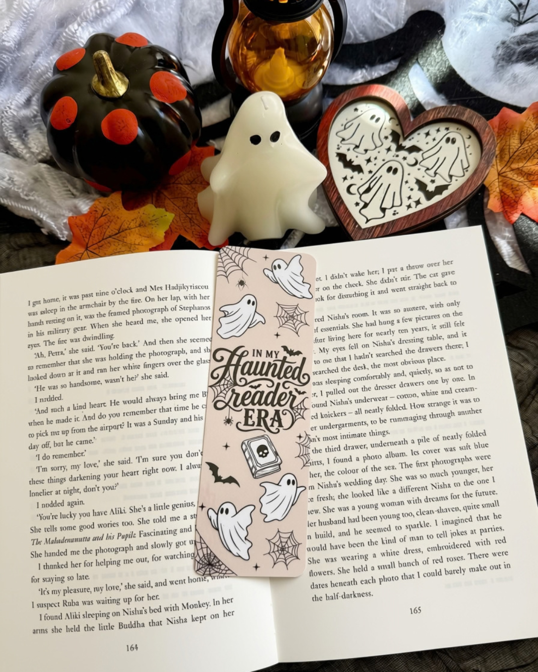 An open printed book spread laid on a gray fabric surface, surrounded by autumn leaves, a white ghost decoration, and a small orange pumpkin. A bookmark is on the page titled "Haunted Reader ERA" with illustrations of a haunted house, spiderwebs, and ghosts.