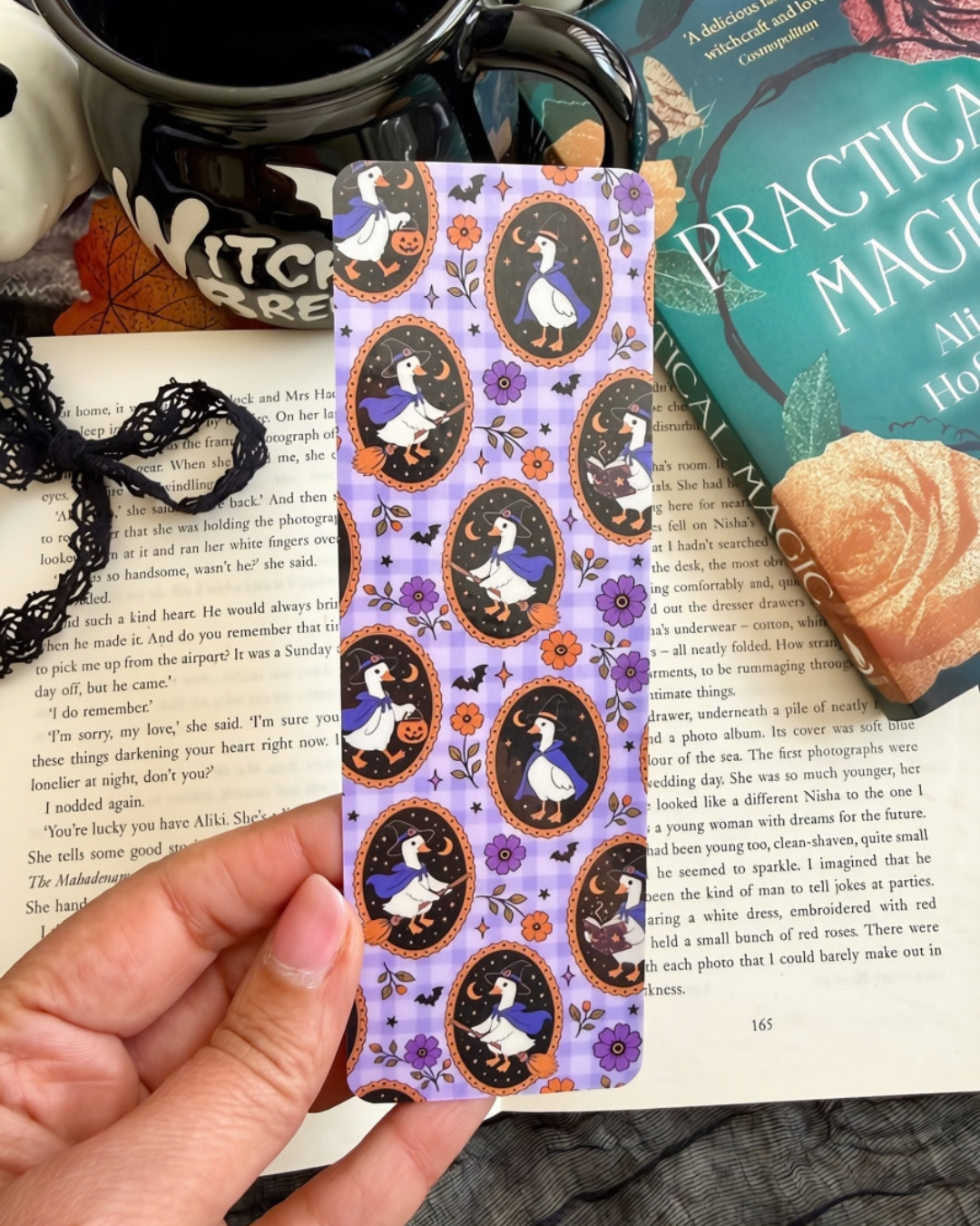 A hand holds a decorative bookmark  featuring a lavender background with repeated oval portraits of a geese in a witches outfits surrounded by orange flowers and purple floral details. The item is photographed in a styled setting with open book pages, black lace, a black teapot, and a green book in the background.