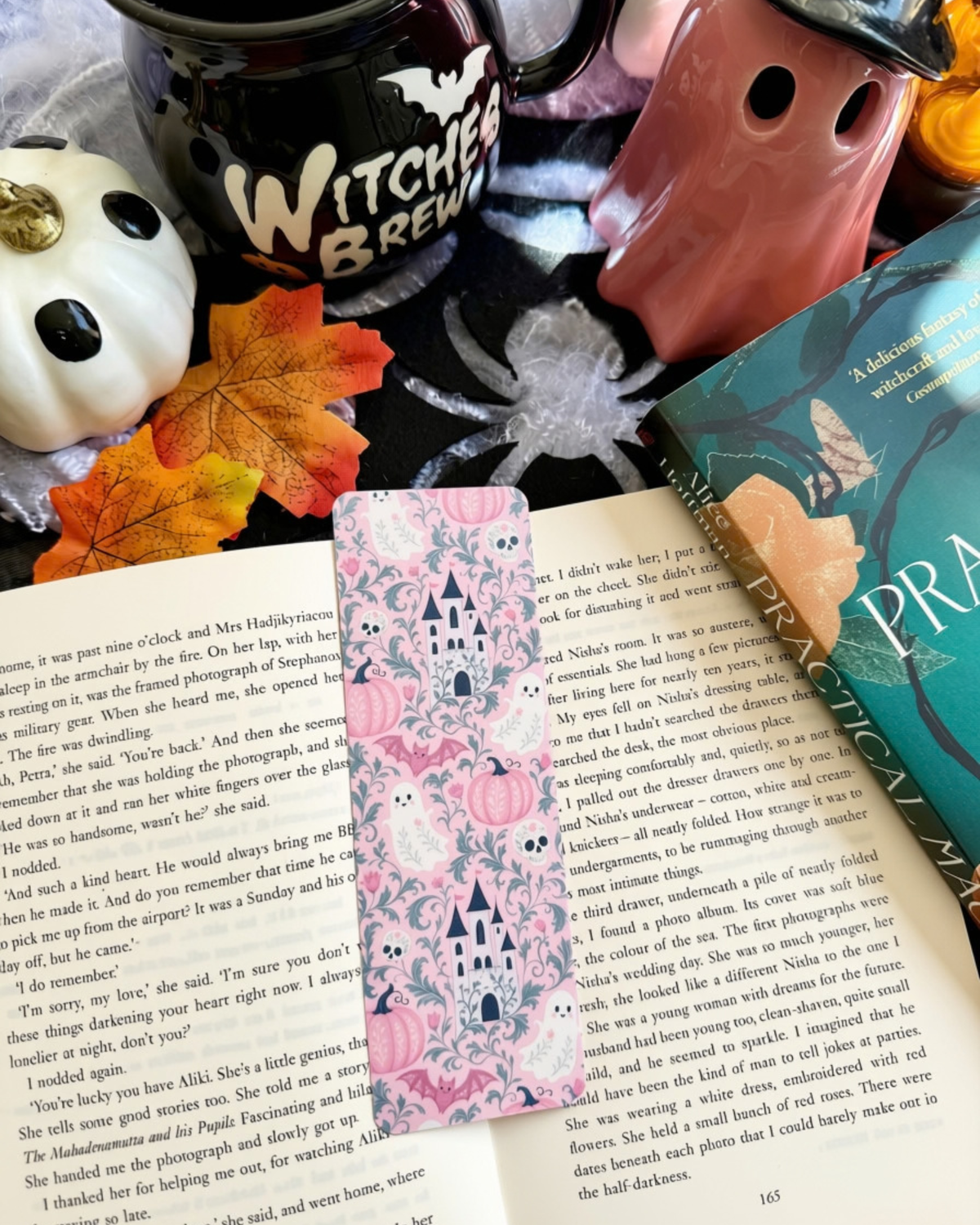 A styled flat-lay featuring a decorative bookmark with pink and green woodland fairy illustrations resting on open book pages, surrounded by autumn leaves, a ceramic mug, and a book.