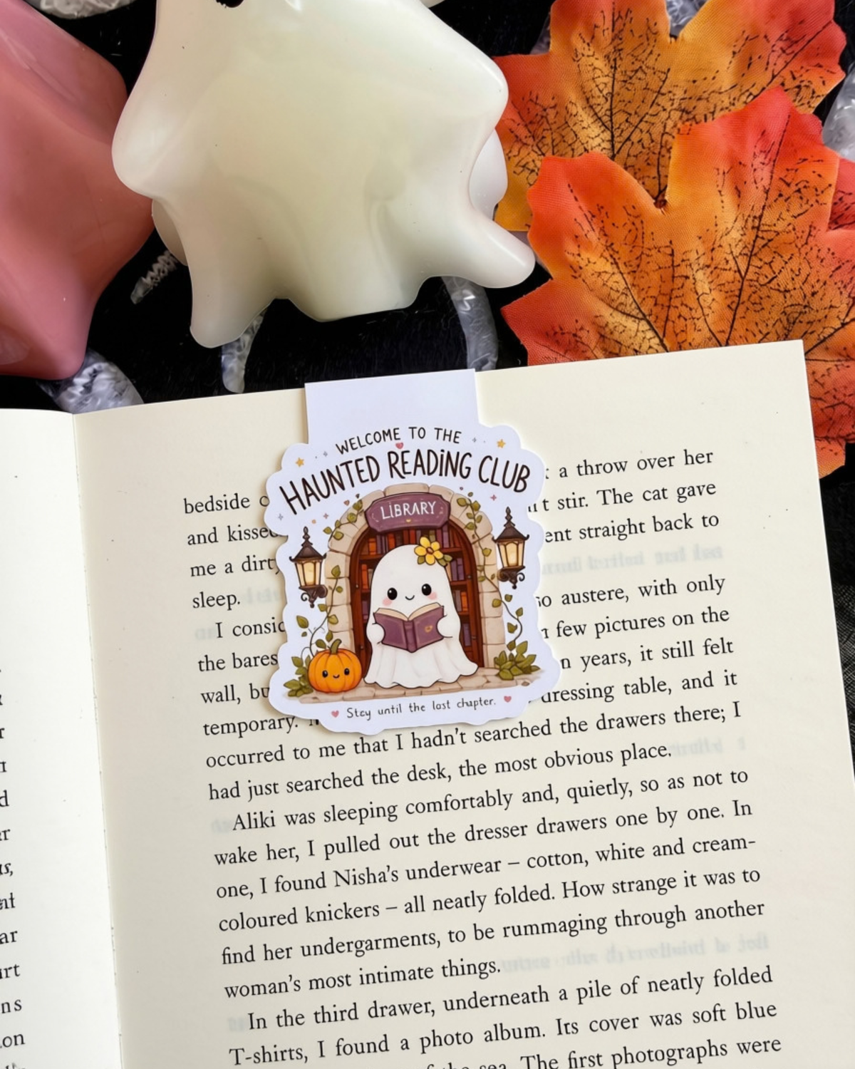 A magnetic bookmark for books, featuring a cartoon haunted house with a library cat and pumpkins, placed on an open book page. The scene is styled with autumn leaves and a ceramic figurine in the background.
