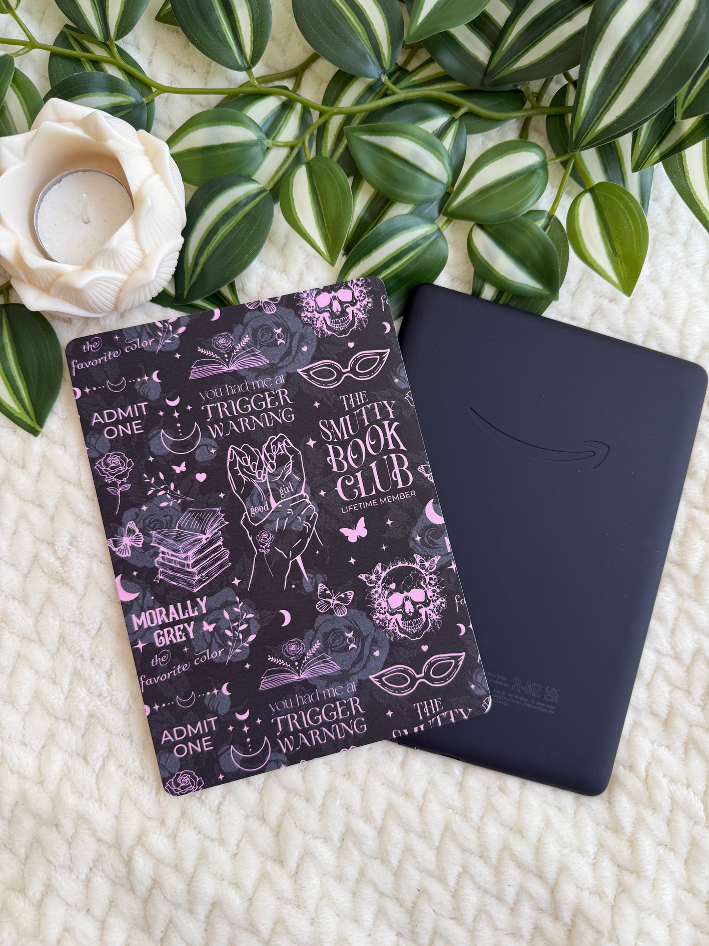 A black Kindle e-reader and a black book cover with pink and white gothic illustrations and text, styled on a cream knit blanket with green leaves and a white rose.