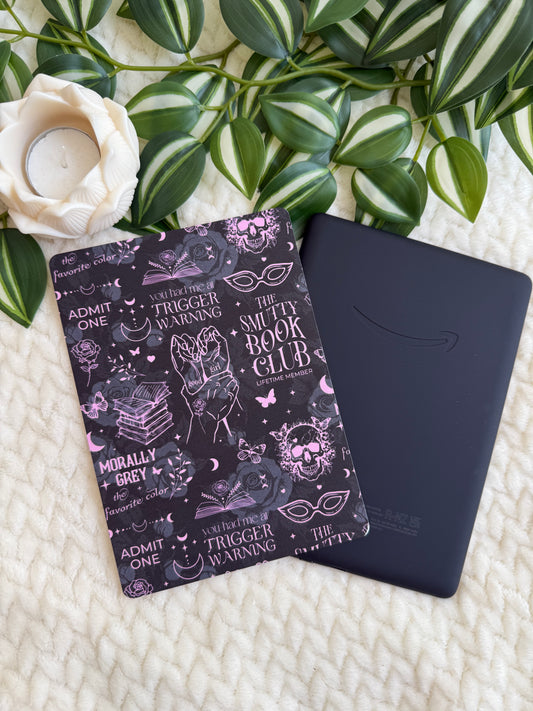 A black Kindle e-reader and a black book cover with pink and white gothic illustrations and text, styled on a cream knit blanket with green leaves and a white rose.