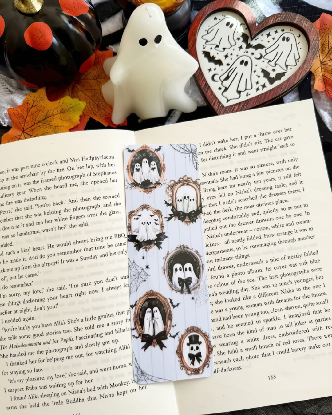 A bookmark with ghost family pics in frames. The bookmark is placed on an open book surrounded by Halloween decorations 