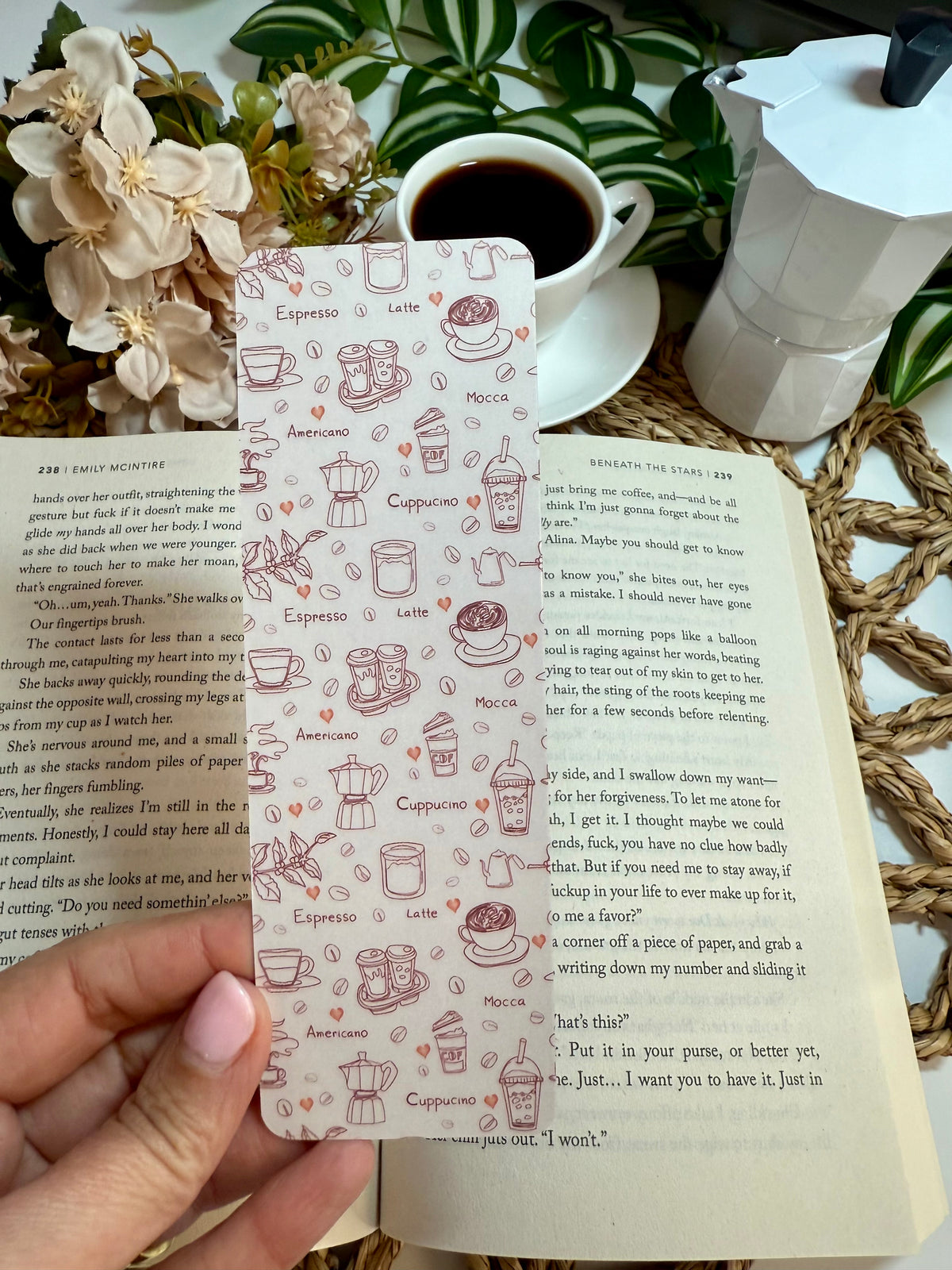 A hand holds a rectangular bookmark featuring delicate brown line drawings of coffee-themed items like espresso cups, latte art, cappuccino, and pastries, set against an open book with other books and a coffee cup in the background.