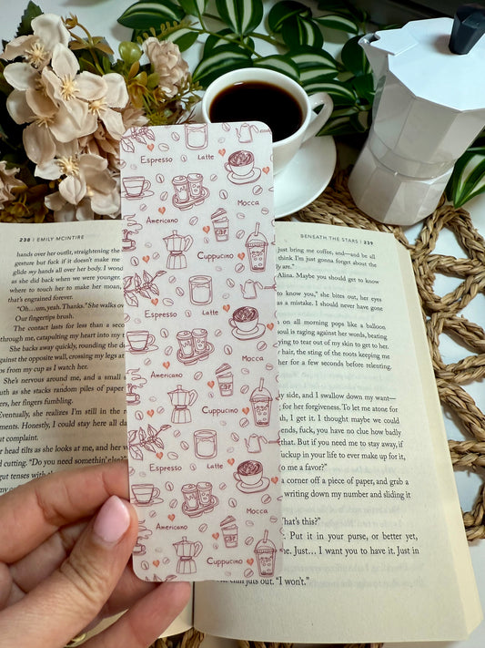 A hand holds a rectangular bookmark featuring delicate brown line drawings of coffee-themed items like espresso cups, latte art, cappuccino, and pastries, set against an open book with other books and a coffee cup in the background.