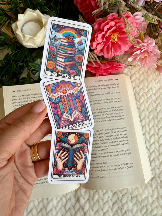 A hand holds a vertical stack of three tarot-style cards titled 'THE BOOK LOVER' over an open book, surrounded by flowers and greenery in a styled setting.