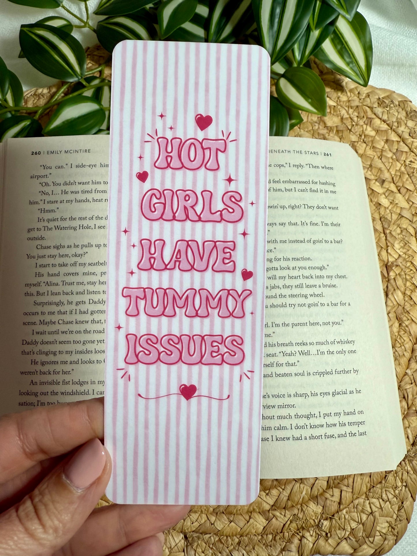 A pink and white vertically striped rectangular card or bookmark with the phrase 'HOT GIRLS HAVE TUMMY ISSUES' in pink lettering and small heart accents, held by a hand over open book pages with green leaves and a woven mat in the background.