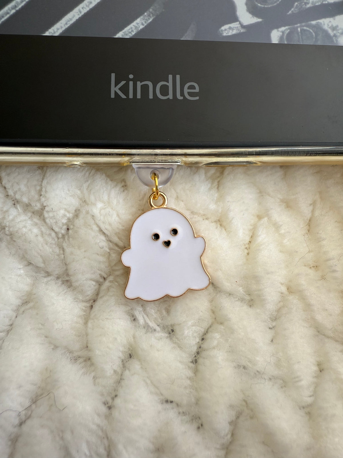 A cute ghost-shaped enamel charm with a gold outline and black eyes, hanging from a clear clip on a gold metal bar. The bar is resting on a white textured knit blanket, with a black Kindle box visible in the background.