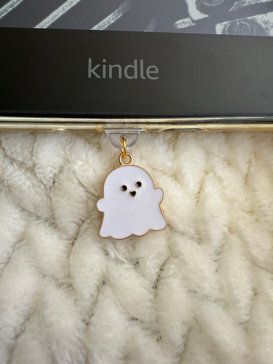 A cute ghost-shaped enamel charm with a gold outline and black eyes, hanging from a clear clip on a gold metal bar. The bar is resting on a white textured knit blanket, with a black Kindle box visible in the background.