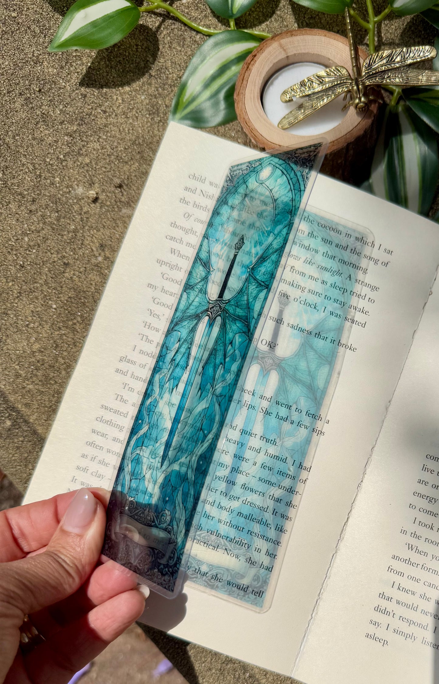 A stained glass bookmark of a sword with dragon wings. The bookmark is placed over an open book allowing the sunlight to filter the image of the bookmark onto the book.