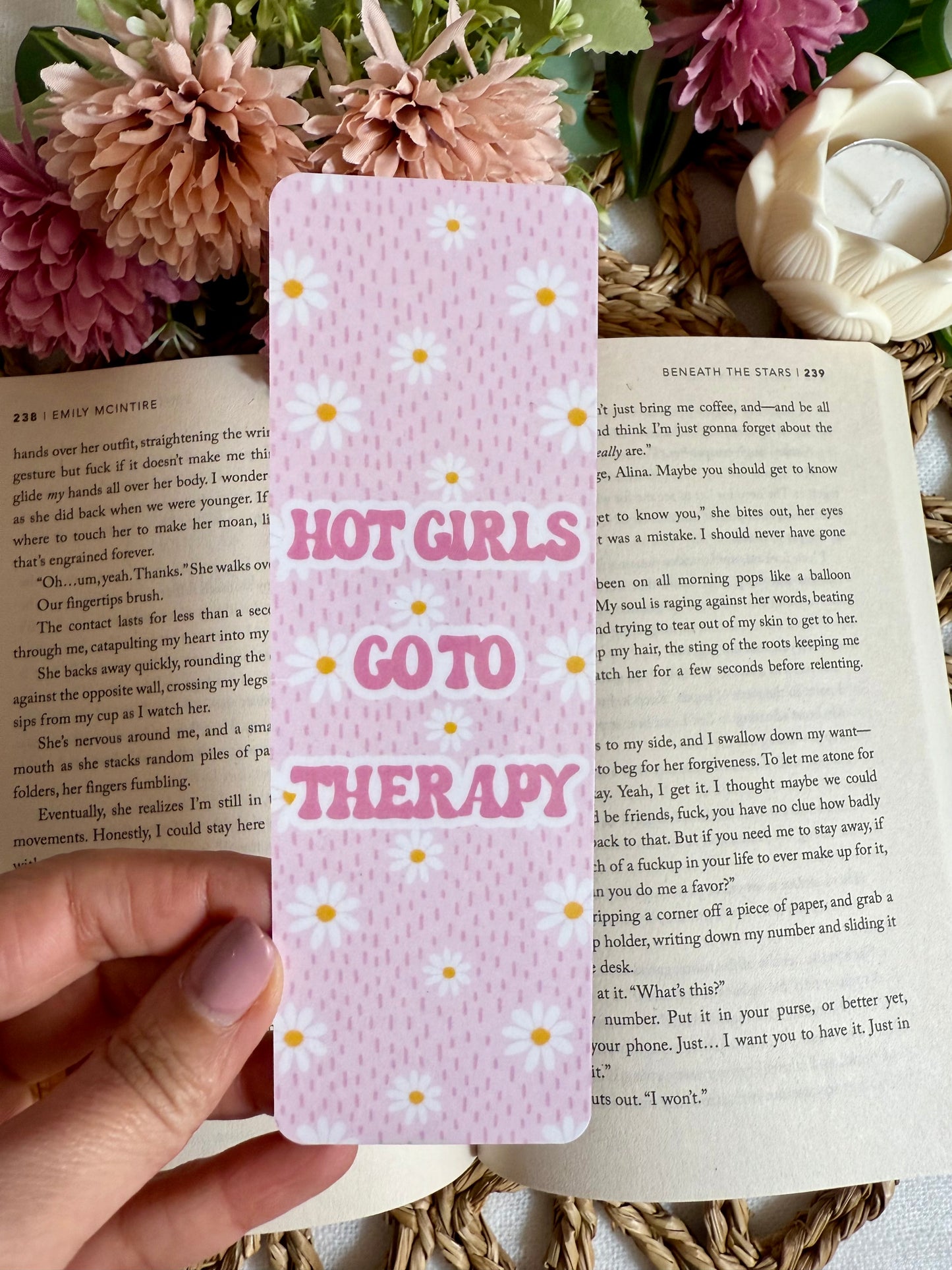 Hand holding a pink bookmark with 'Hot Girls Go to Therapy' text, surrounded by flowers and a shell.
