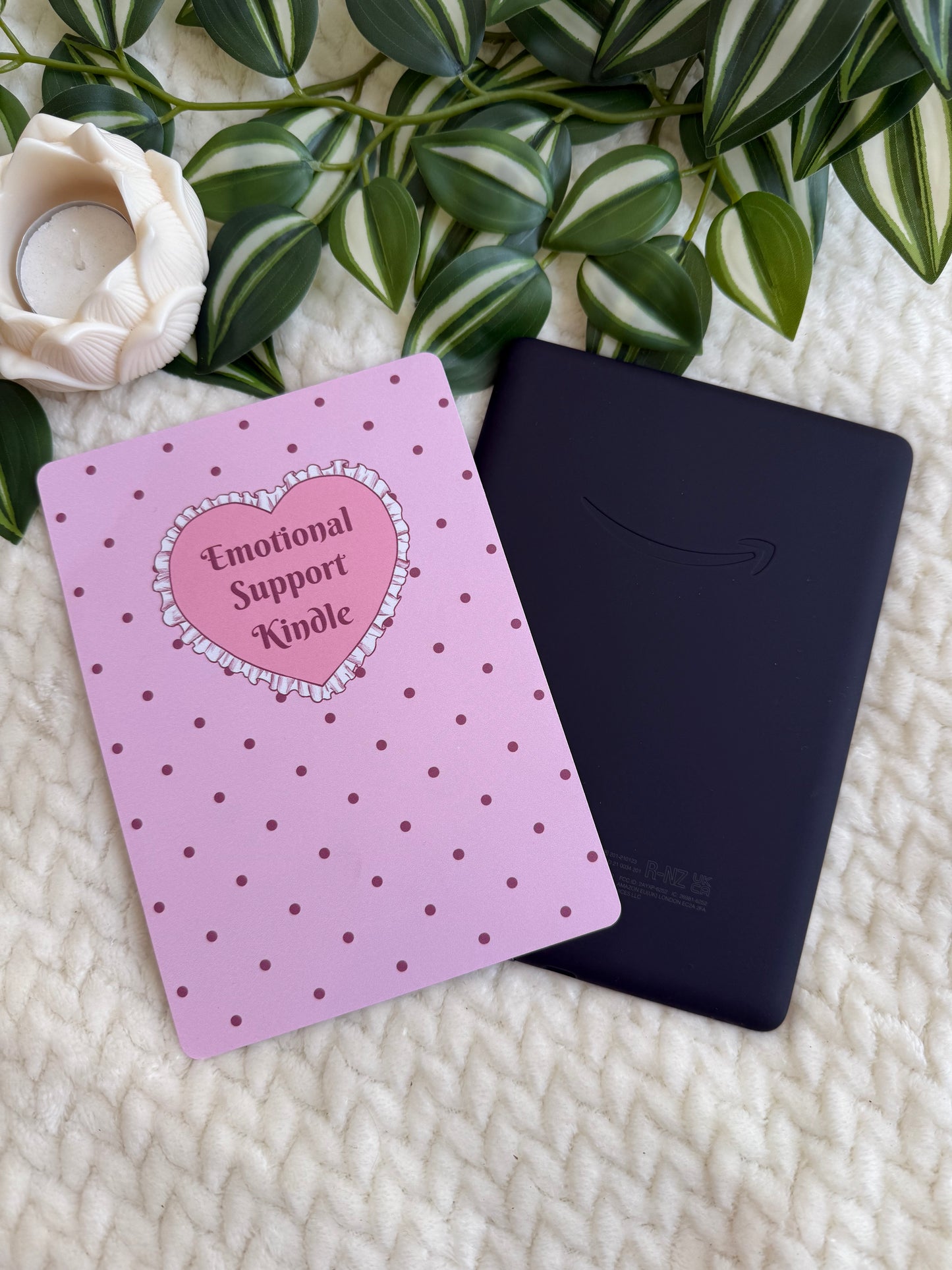 A pink Kindle e-reader with a heart-shaped 'Emotional Support Kindle' sticker on the front, placed next to a black Kindle device on a textured cream blanket with green leaves and a white rose in the background.