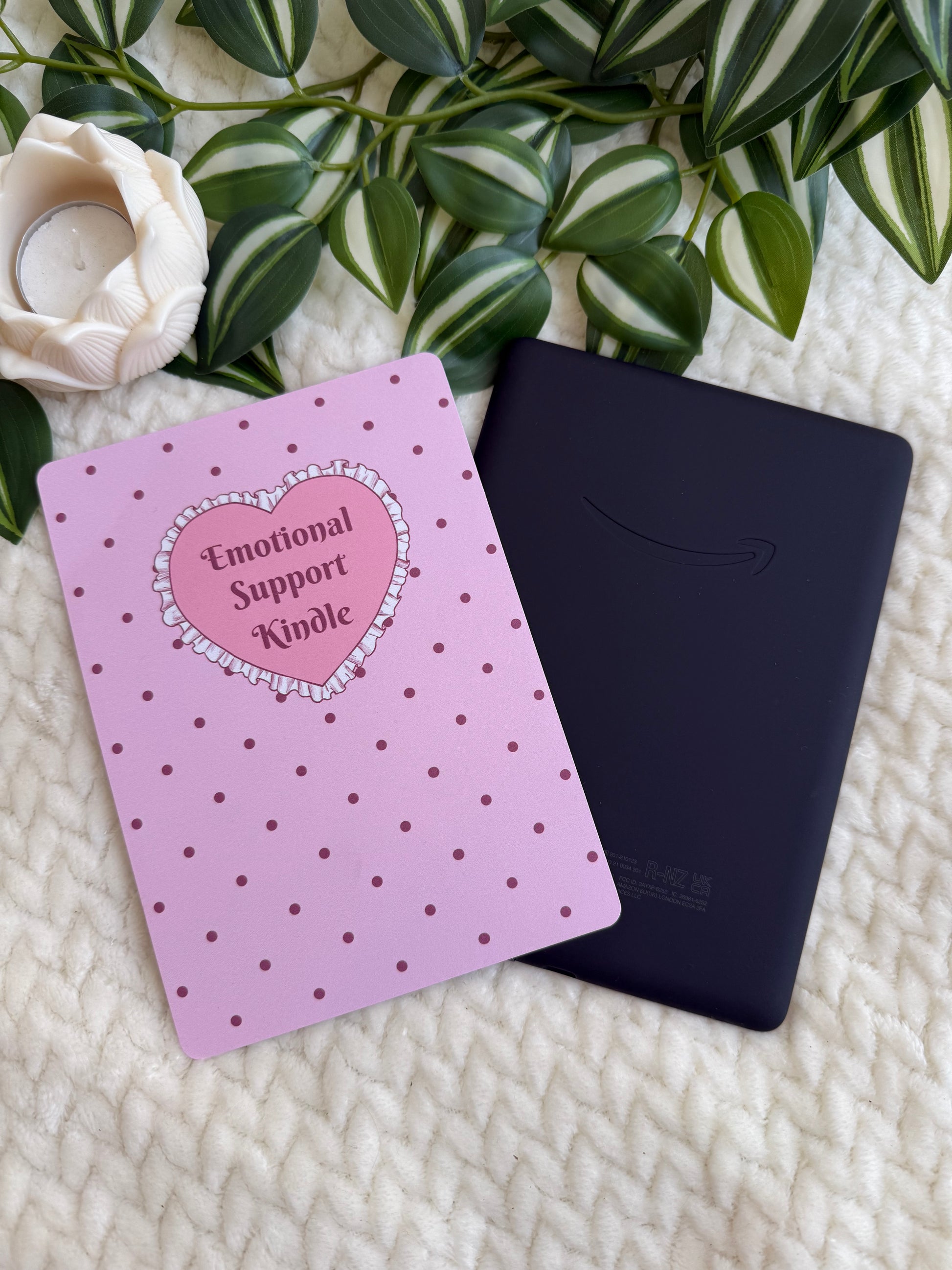 A pink Kindle e-reader with a heart-shaped 'Emotional Support Kindle' sticker on the front, placed next to a black Kindle device on a textured cream blanket with green leaves and a white rose in the background.