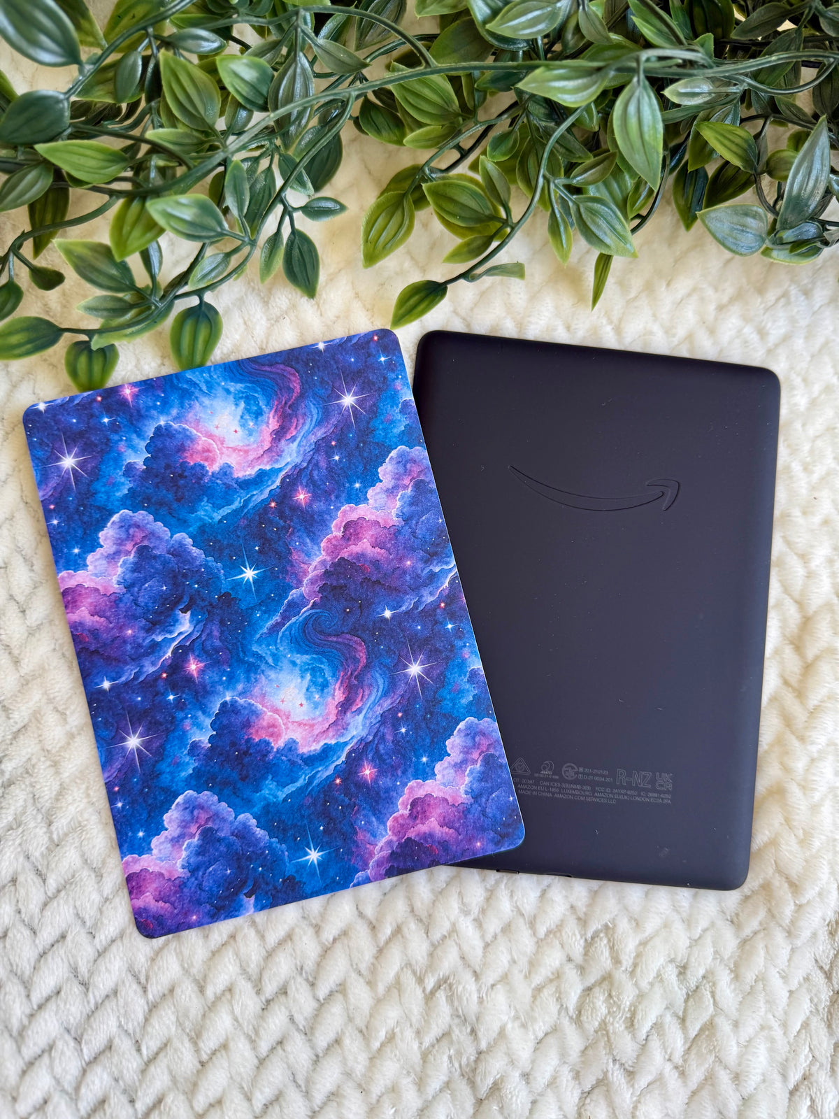 A black Kindle e-reader device with the Amazon smile logo lies next to a colorful galaxy-patterned sleeve or cover on a cream textured blanket with green leaves at the top.