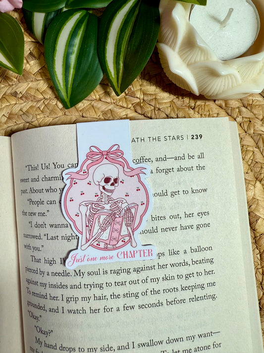 A decorative bookmark featuring a pink ribbon and a skeleton in a dress with a bow, clipped to an open book. The bookmark is styled on a woven mat with green leaves and a white ceramic dish in the background.