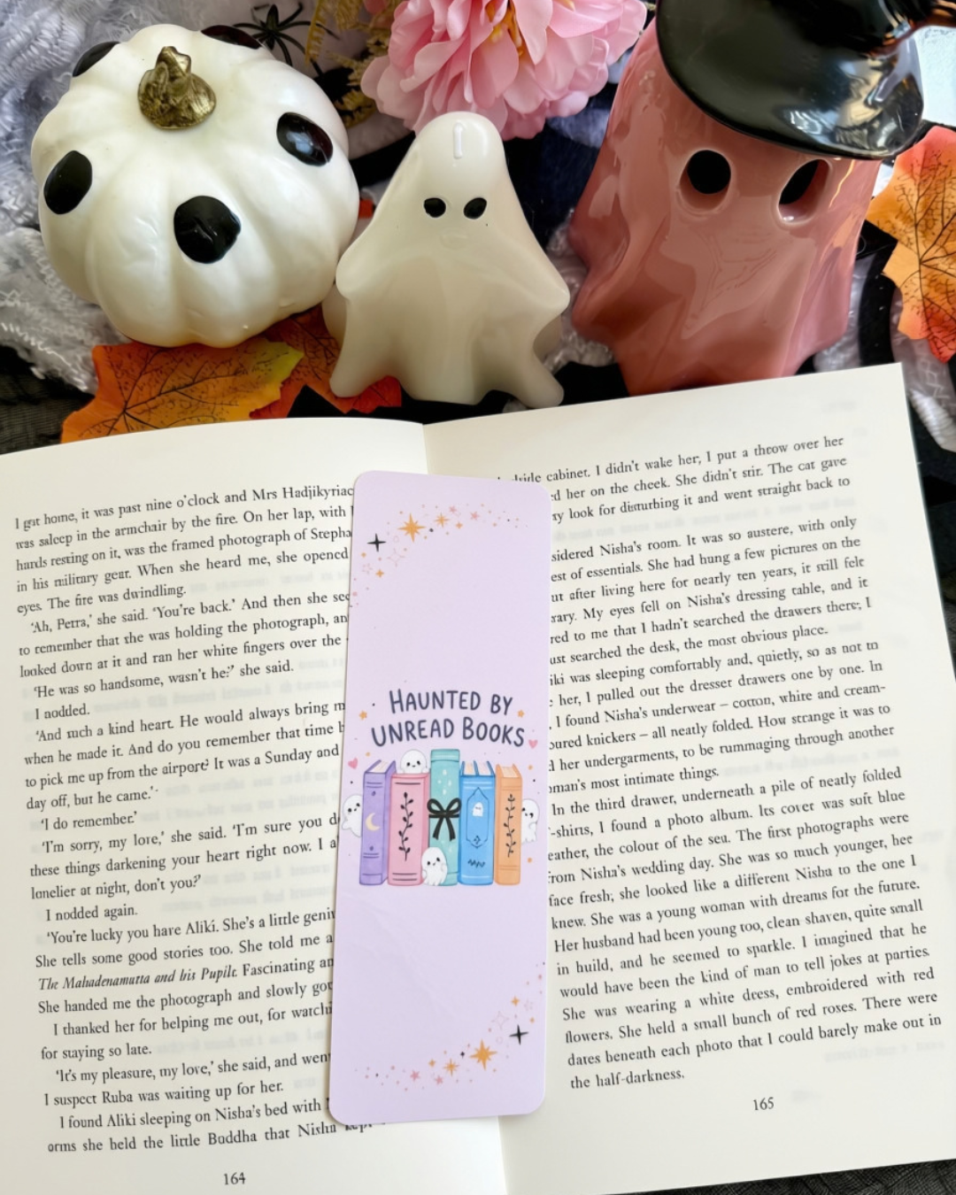 A decorative bookmark card with the text 'HAUNTED BY UNREAD BOOKS' and illustrations of a purple book and a pink book, placed on an open book between two white ceramic dog toys with pink flowers.