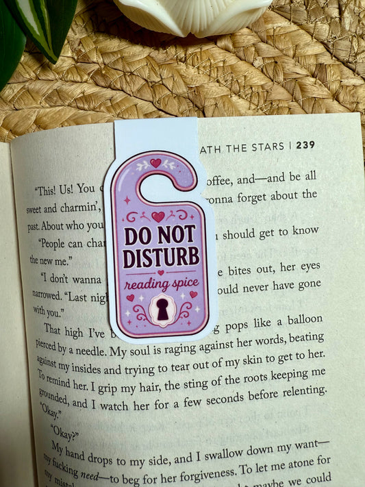 A decorative magnetic bookmark shaped like a hanging doorbell in lavender and pink, printed with the text 'DO NOT DISTURB reading spice,' placed on an open book page.
