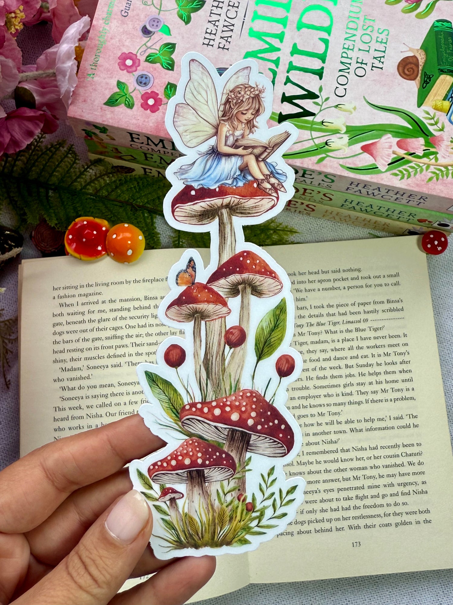 A die cut bookmark of a Fairy sitting on a stack of mushrooms reading a book, with whimsical cosy fantasy books in the background