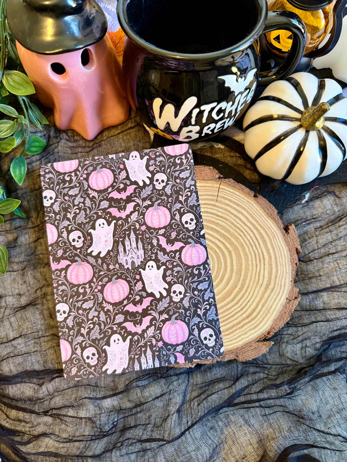 A kindle insert with a Halloween pattern of pink pumpkins, white ghosts, and black skulls on a dark background, displayed on a wood slice with witch-themed props including a black mug, a ghost-shaped container, and a small white pumpkin.