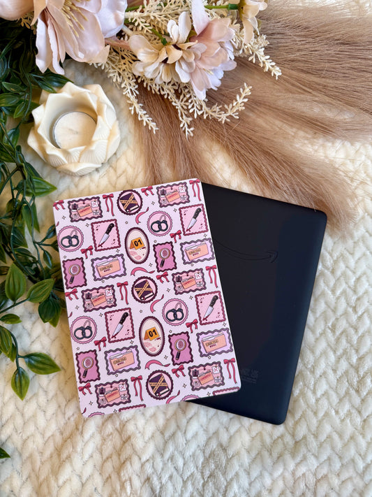 A black tablet or e-reader device placed on top of a pink patterned insert with thriller themed illustrations, styled on a cream knit blanket with flowers, greenery, and faux fur.