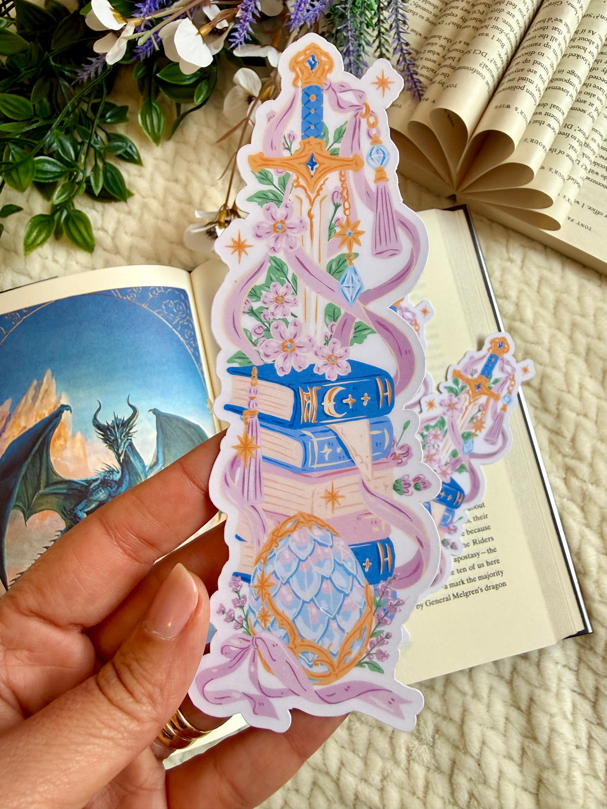 A die cut bookmark of a fantasy sword in pile of books. the bookmark is placed over an open book with a page of a dragon