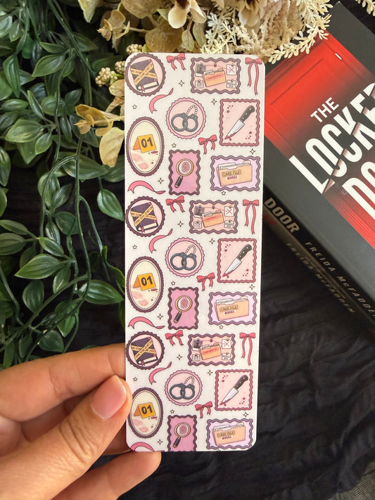 A hand holds a thriller themed bookmark featuring a repeating pattern of pink and purple crime scene investigation themed stamps, magnifying glasses, and evidence bags, with a '5' inside a badge. The background features a black fabric backdrop, green foliage, dried flowers, and a book spine.