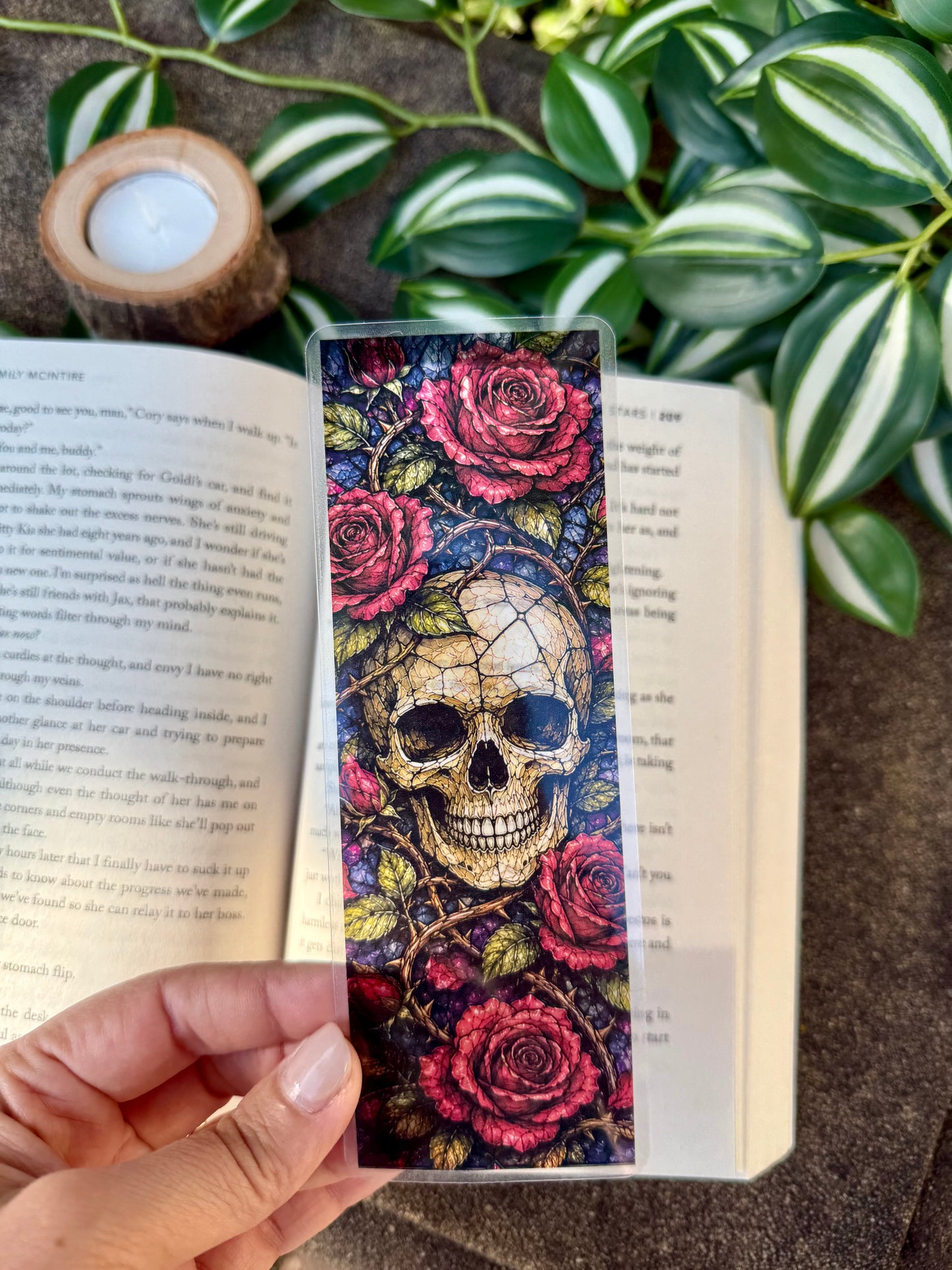 Colorful stained glass skull and rose bookmark held over an open book with plants in the background